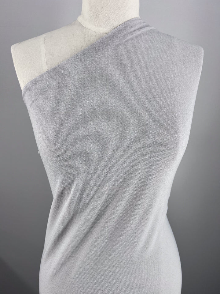 A mannequin wears a one-shoulder dress made from Super Cheap Fabrics' Crepe Lycra in Smoke. This medium weight jersey fabric drapes elegantly, its simple design contrasting beautifully against a plain grey background.