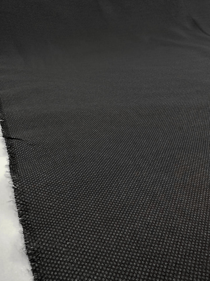 Close-up of "Printed Bengaline - Pin Dot Black" by Super Cheap Fabrics, showcasing a dark gray, medium-weight fabric with a grid-like texture. Ideal for skirts and dresses, it stretches diagonally with frayed edges on the left. The surface reflects a subtle sheen under soft lighting.