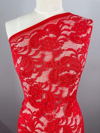 A mannequin displays Super Cheap Fabrics' Designer Lace - Scarlet - 145cm, showcasing its vibrant scarlet hue and intricate floral pattern. The sheer lace, perfect for evening dresses, is styled asymmetrically over one shoulder for an elegant look.