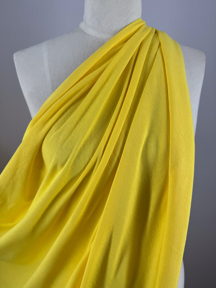 A Super Cheap Fabrics mannequin is draped with Cotton Jersey - Golden Kiwi - 155cm, flowing smoothly over the shoulder and creating soft folds. The neutral gray backdrop highlights the vibrant color and texture of this fabric.