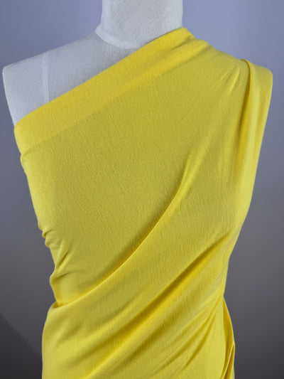 A dressmaker's mannequin features a striking one-shoulder design using Super Cheap Fabrics' Golden Kiwi fabric. The Cotton Jersey - Golden Kiwi - 155cm enhances the graceful flow against the plain grayish-blue background.