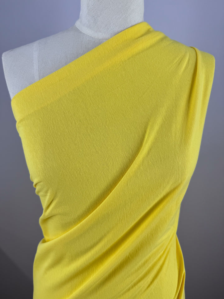 A dressmaker's mannequin features a striking one-shoulder design using Super Cheap Fabrics' Golden Kiwi fabric. The Cotton Jersey - Golden Kiwi - 155cm enhances the graceful flow against the plain grayish-blue background.