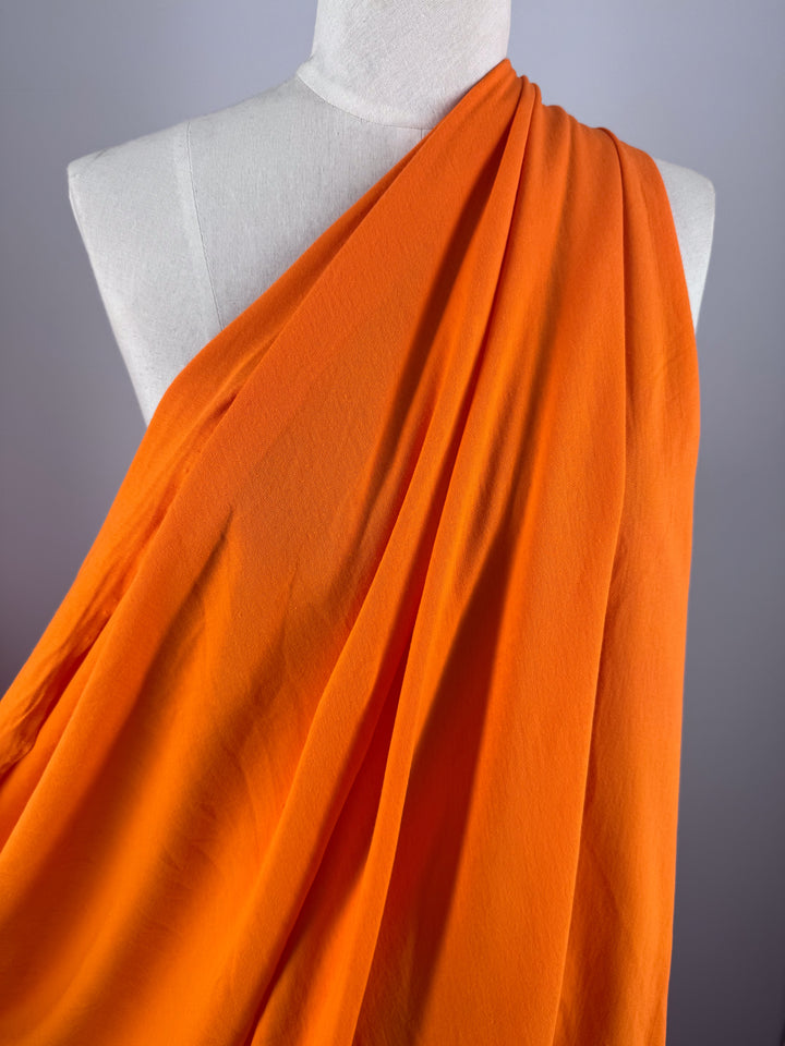 A close-up of the "Cotton Jersey - Vibrant Orange - 155cm" by Super Cheap Fabrics showcases lightweight, elegantly draped fabric over a mannequin, with soft folds and dynamic movement. The neutral backdrop accentuates its rich color and texture.