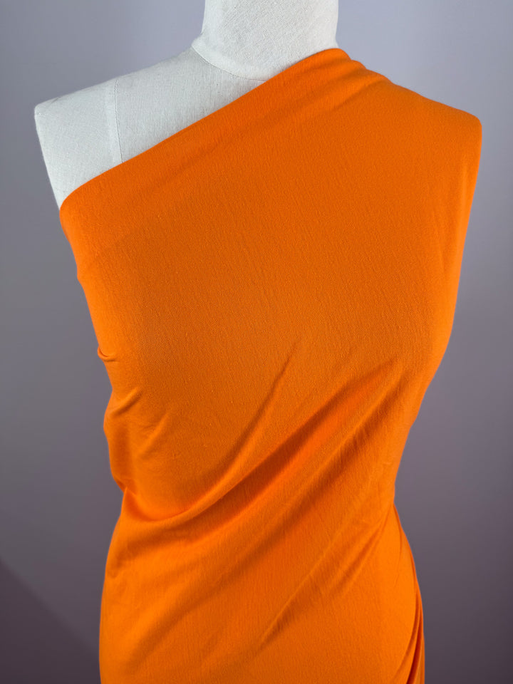 A mannequin showcases Super Cheap Fabrics' Cotton Jersey in Vibrant Orange (155cm), elegantly draped over one shoulder, forming a diagonal line across the chest. The smooth fabric's subtle creases contrast with the light gray backdrop.