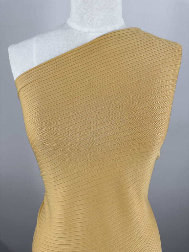 A mannequin showcases the Rib Knit - Pale Golden Nugget by Super Cheap Fabrics, featuring a one-shoulder design against a neutral backdrop. This 173cm medium-weight fabric has horizontal ribbing that lends a structured look.