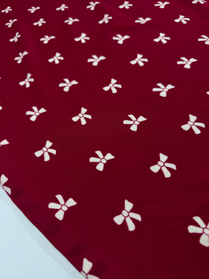 The Printed Cotton Lycra - Bow from Super Cheap Fabrics, in an Urban Red hue, displays evenly spread small white bow-like patterns across its smooth, slightly shiny surface.