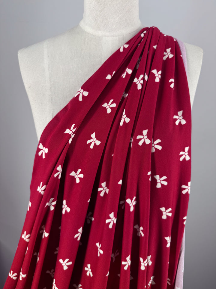 A mannequin displays the "Printed Cotton Lycra - Bow - 150cm" by Super Cheap Fabrics, featuring small white bows on a deep Urban Red background of medium-weight lycra fabric.
