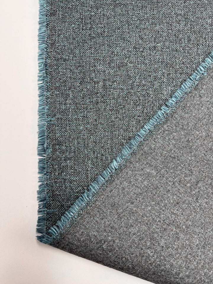 A close-up of Super Cheap Fabrics' Upholstery - Soho Topaz-150cm shows textured gray fabric with blue fringed edges, partially folded to reveal both sides, set against a light background.