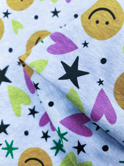 The Printed Jersey - Hallow from Super Cheap Fabrics features a lightweight fabric with playful patterns of yellow smiley faces, pink and green hearts, and black stars on a light background, perfect for creative projects.