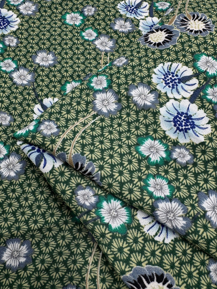 The Printed Crepe - Botan from Super Cheap Fabrics features elegant green fabric with detailed geometric patterns and floral designs in blue, white, and black. The flowers are intricate and scattered across the 148cm textile, which appears layered.