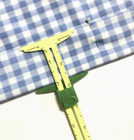 A Sliding Sewing Gauge from Super Cheap Fabrics, resting on blue and white checkered fabric with a marking pin, is ready to ensure precision. The gauge, marked in centimeters, meticulously measures the seam allowance for perfect quilting.
