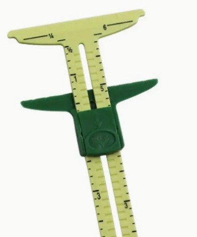 The Sliding Sewing Gauge by Super Cheap Fabrics is a green tool for measuring knitting or sewing dimensions. It features vertical and horizontal scales with numbers and fractions, making it essential for precise quilting adjustments.