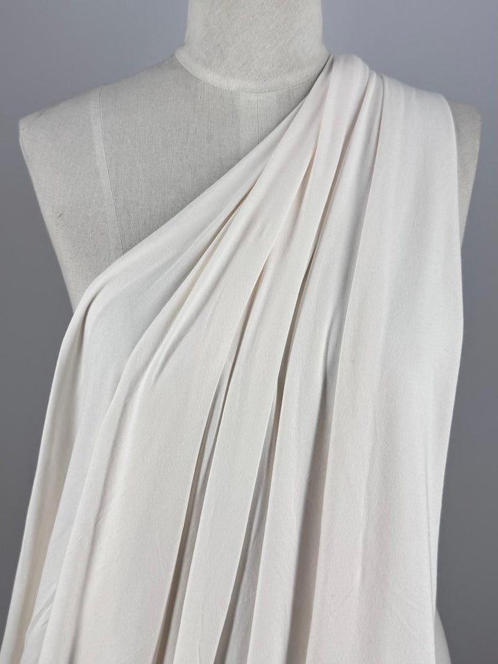 A dress form showcases Super Cheap Fabrics' Cotton Lycra - Sugar Swizzle (135cm), a medium weight cream-colored fabric. Its toga-like draped elegance flows over one shoulder against a plain gray backdrop, accentuating the stretch and folds of the material.