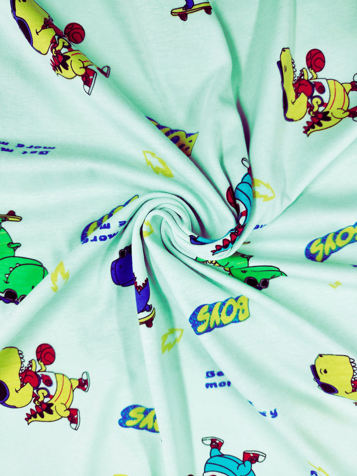 The Super Cheap Fabrics "Printed Jersey - Dino - 155cm" features a light green fabric with cartoon dogs playing basketball and skateboarding on a spiral pattern. It includes playful "SKO" and "Boys" words in comic style, ideal for comfortable childrenswear with its two-way stretch.