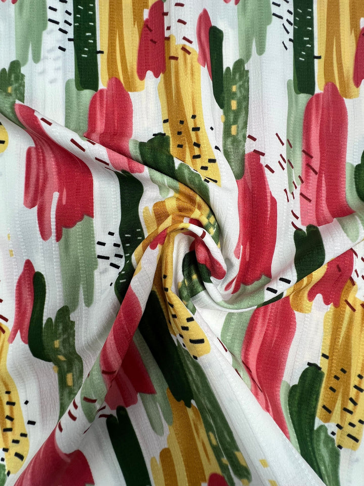 A close-up of Super Cheap Fabrics' Textured Deluxe Print - Stroke & Speck 2.0 (155cm), featuring an abstract pattern with vertical brushstrokes in red, green, yellow, pink, and black/yellow dashed lines on white polyester, loosely twisted at center.