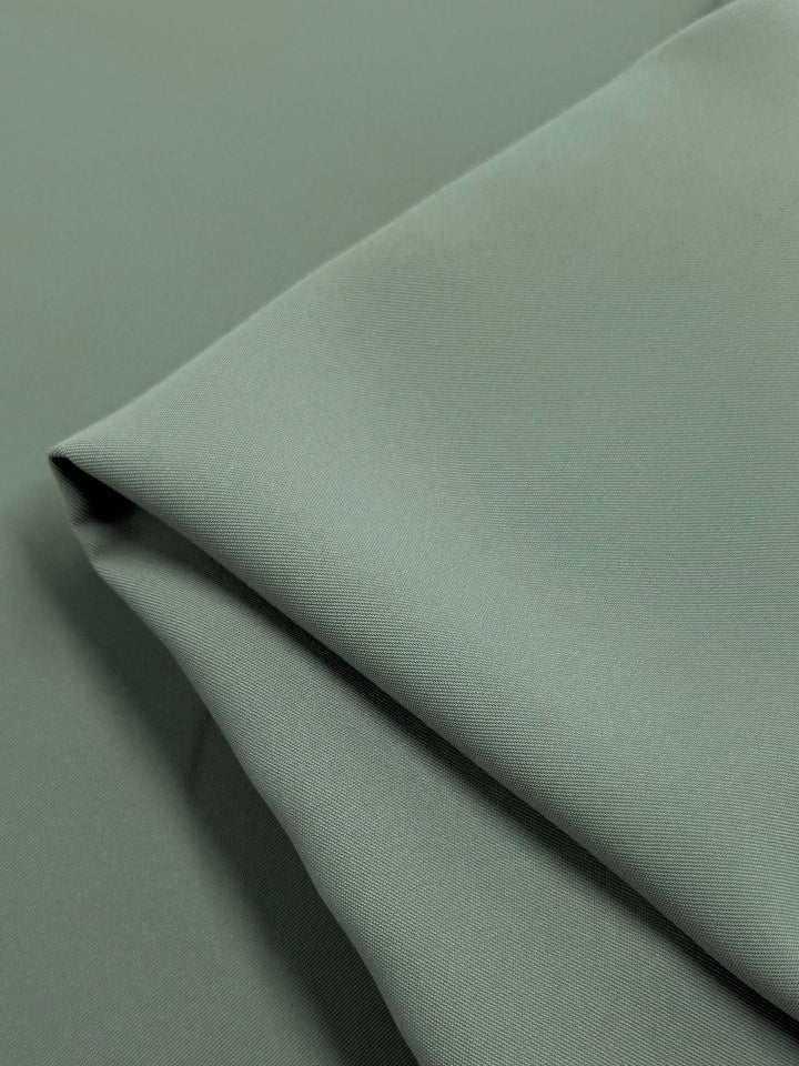 A detailed view of Super Cheap Fabrics' Microfibre Suiting in Dried Sage shows the medium-weight, light green fabric's subtly textured surface. The 150cm polyester appears soft and tidy, with clean lines and gentle shadows highlighting its elegant folds.