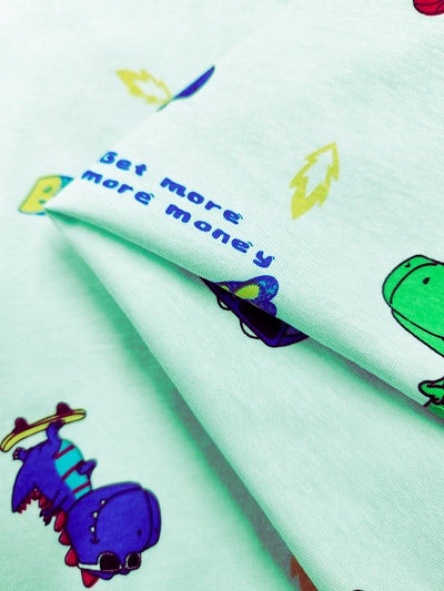 Close-up of Super Cheap Fabrics' Dino Printed Jersey, a light green childrenswear fabric with cheerful dinosaur illustrations, including a purple-and-green dino. "Get more, more money" is in blue surrounded by playful yellow leaves. Made from 155cm wide jersey with two-way stretch for comfort and fun.