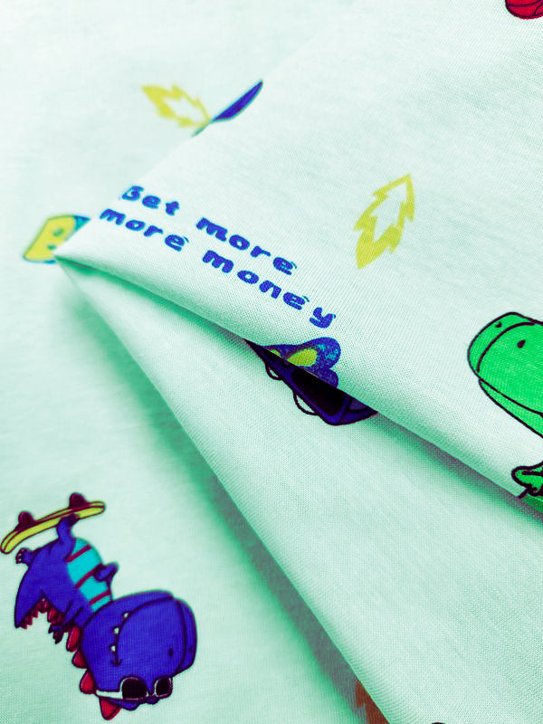 Close-up of Super Cheap Fabrics' Dino Printed Jersey, a light green childrenswear fabric with cheerful dinosaur illustrations, including a purple-and-green dino. "Get more, more money" is in blue surrounded by playful yellow leaves. Made from 155cm wide jersey with two-way stretch for comfort and fun.