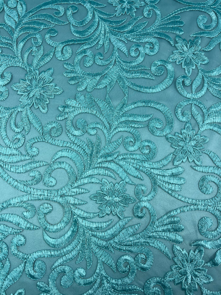 A close-up of an intricate teal fabric featuring an embroidered floral and swirling vine pattern. The threads are slightly shiny, creating a textured and detailed Evening Lace - Aqua Galadriel - 125cm design against the lighter teal background from Super Cheap Fabrics.