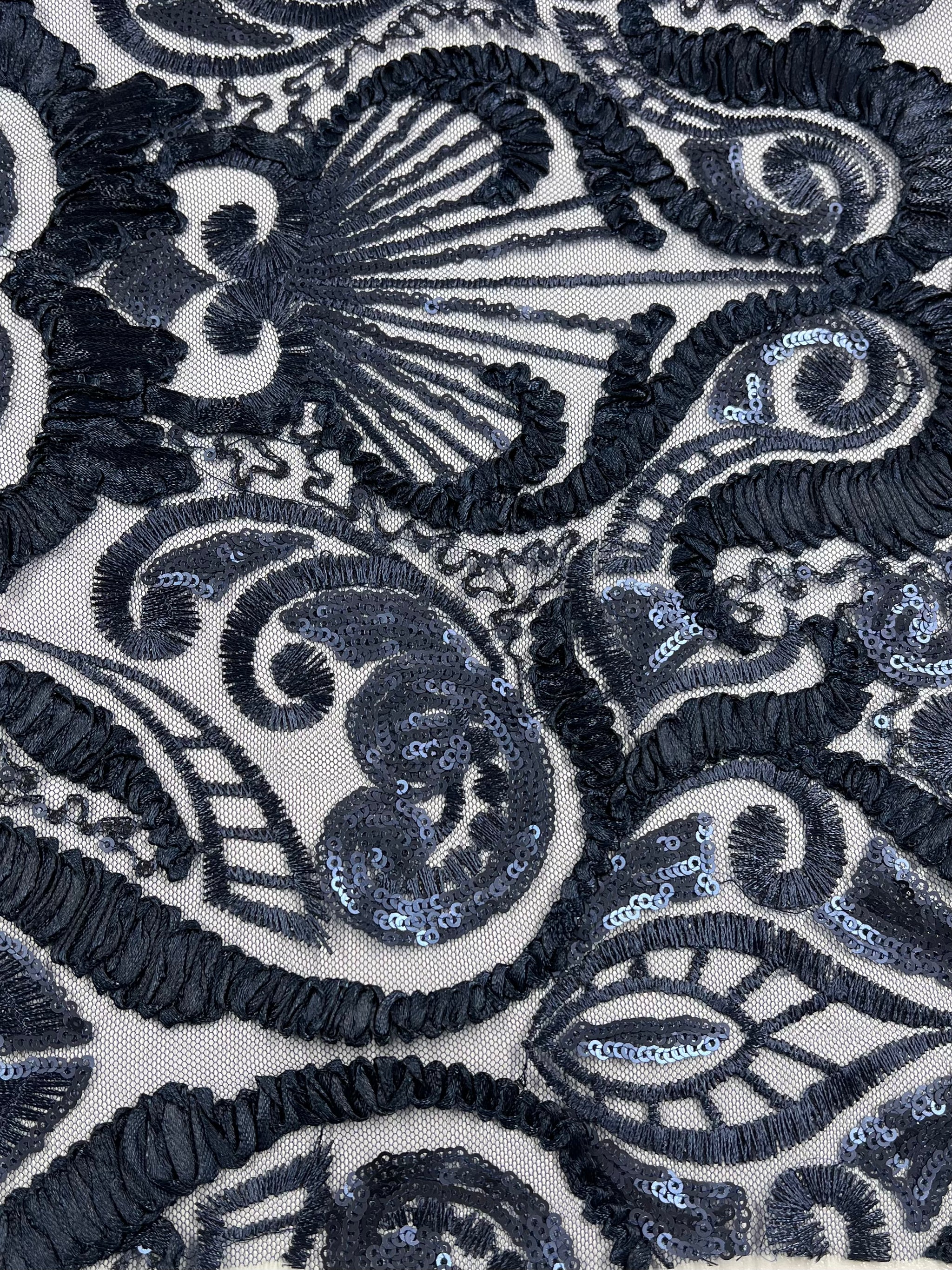 Close-up view of an intricate black lace fabric with an ornate pattern. The medium weight fabric features a combination of floral and swirling designs adorned with shimmering sequins, giving it a textured and elegant appearance. The sheer base, made from 100% polyester, enhances the detailed design. This is the 