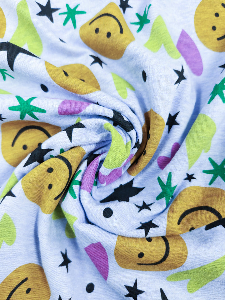 A close-up of Super Cheap Fabrics' Printed Jersey - Hallow, 165cm, reveals a lightweight fabric with yellow smiley faces, black stars, and green and purple abstract shapes on a light gray background.