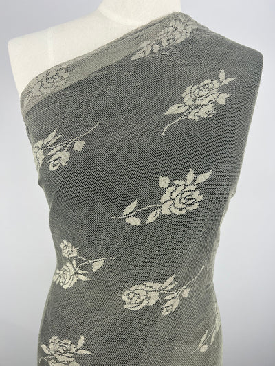 The mannequin is draped with Super Cheap Fabrics' Floral Lace in Olive, measuring 125cm. This lightweight polyester features intricate white rose patterns on a textured gray background, enhancing its stylish contrast, perfect for fashionable tops.