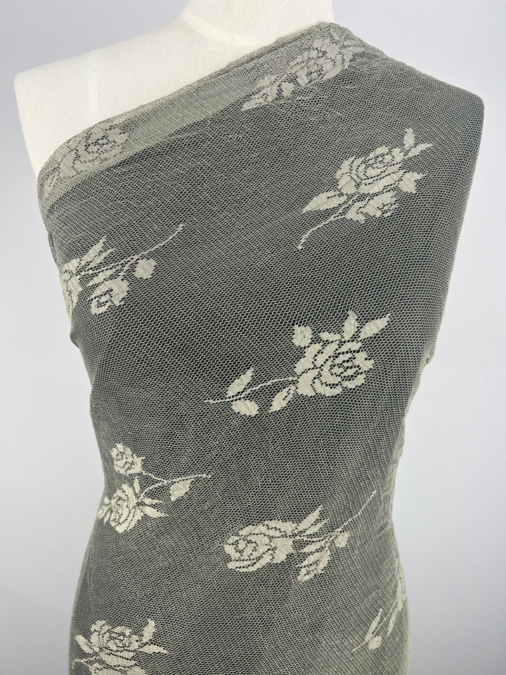 The mannequin is draped with Super Cheap Fabrics' Floral Lace in Olive, measuring 125cm. This lightweight polyester features intricate white rose patterns on a textured gray background, enhancing its stylish contrast, perfect for fashionable tops.