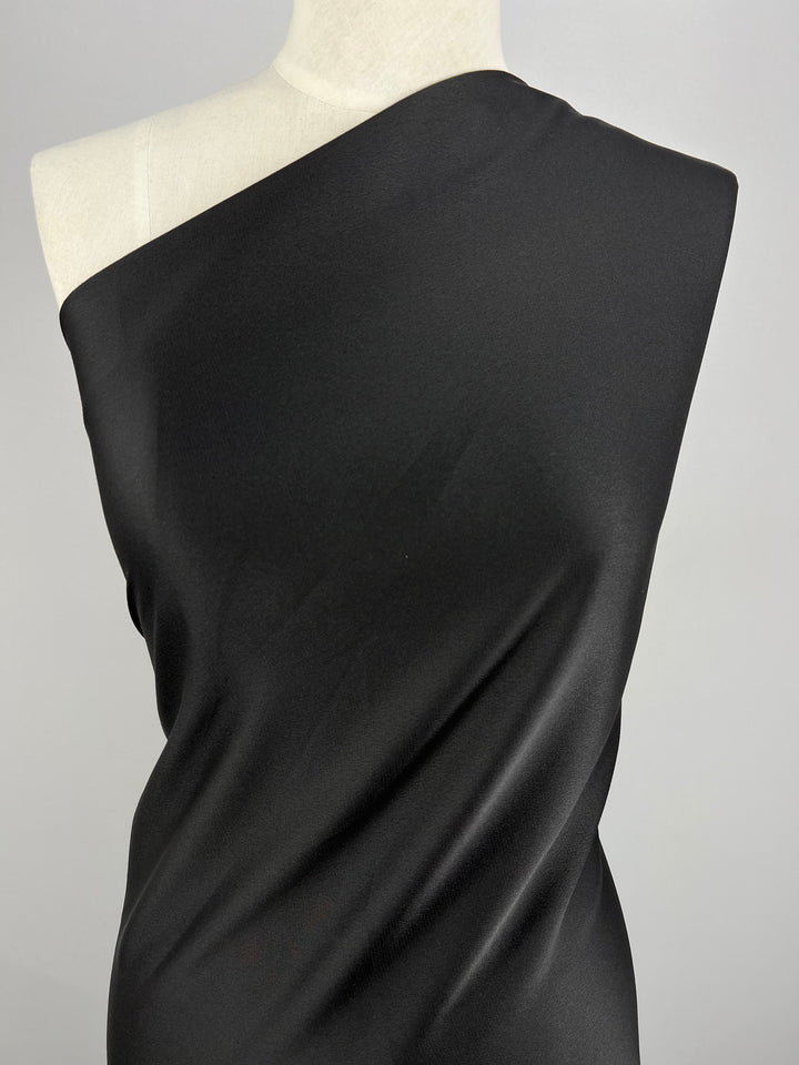A one-shoulder black dress draped on a mannequin against a plain white background. The medium weight Crepe De Chine - Black - 150cm fabric from Super Cheap Fabrics appears smooth and slightly shiny, creating elegant folds that enhance the dress's sleek and sophisticated look.