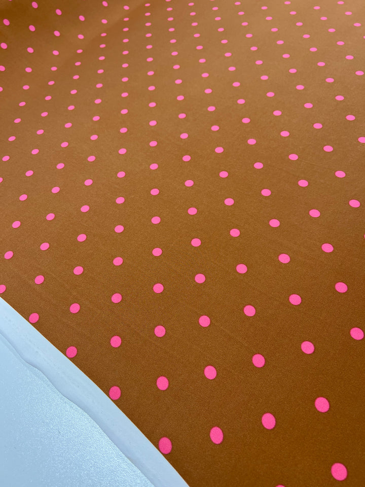 A close-up of Super Cheap Fabrics' Printed Lycra - Pecan & Pink Polka (150cm), a soft medium-weight fabric featuring evenly spaced small pink polka dots on brown with a white border along the bottom left edge.