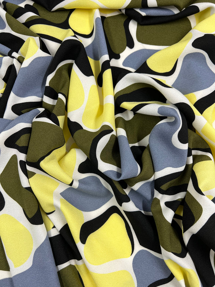 The "Printed Crepe - Camo Special - 150cm" by Super Cheap Fabrics showcases a vibrant, lightweight polyester fabric with an abstract pattern of interlocking shapes in yellow, black, white, olive green, and gray. It appears soft and draped with folds and shadows, ideal for versatile uses.