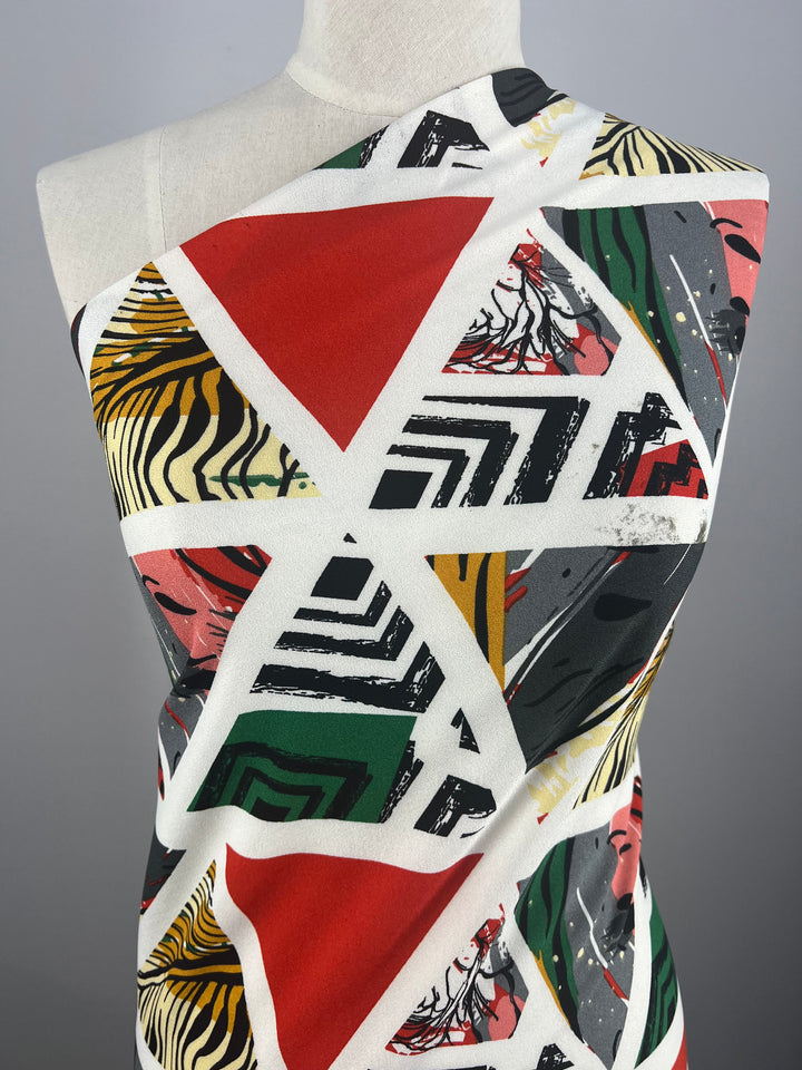 The Printed Crepe - Windows by Super Cheap Fabrics, draped on a mannequin, displays a lightweight polyester with geometric patterns including triangles and abstract designs in a multi-color palette of red, black, green, yellow, and white featuring stripes and organic shapes. Width: 150cm.