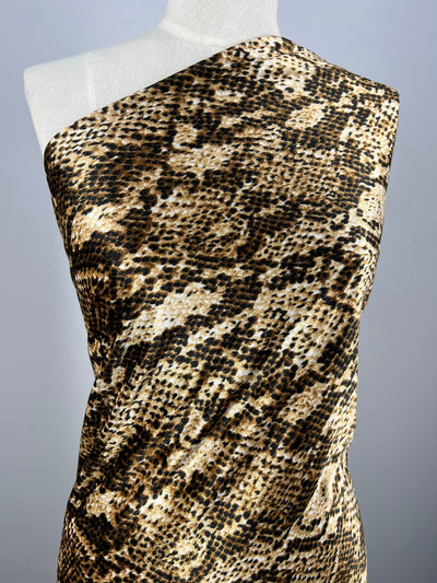 A white mannequin displays a one-shoulder dress in Super Cheap Fabrics’ Printed Velvet - Python, a light to medium weight polyester with a brown, black, and beige snakeskin pattern. The fabric is 150cm wide. Background is plain and neutral.