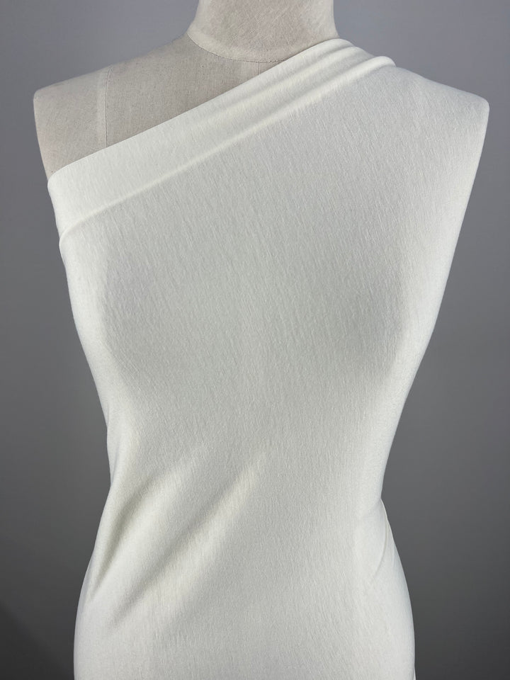 A minimalist, off-the-shoulder ivory dress on a mannequin, draped elegantly with a single shoulder neckline. The medium weight Cotton Lycra - Ivory - 150cm from Super Cheap Fabrics appears soft and smoothly fitted, highlighting the simplicity and elegance of the design against a plain gray background.