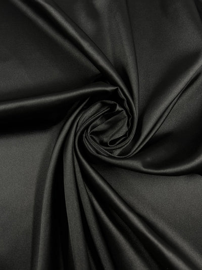 A close-up image of smooth, black **Satin Deluxe - Black - 150cm** fabric from **Super Cheap Fabrics**, with a portion twisted into a spiral pattern in the center. The material has a glossy sheen, reflecting light off the surface, highlighting its luxurious texture. Ideal for evening wear fabric.