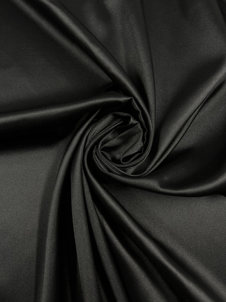 A close-up image of smooth, black **Satin Deluxe - Black - 150cm** fabric from **Super Cheap Fabrics**, with a portion twisted into a spiral pattern in the center. The material has a glossy sheen, reflecting light off the surface, highlighting its luxurious texture. Ideal for evening wear fabric.